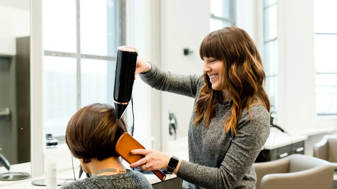 Een kapster glimlacht terwijl ze het haar van een klant föhnt in een lichte salon