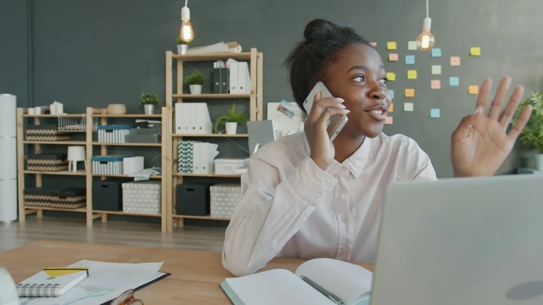 Een lachende vrouw aan de telefoon aan haar bureau, met een laptop en memoblaadjes op de muur